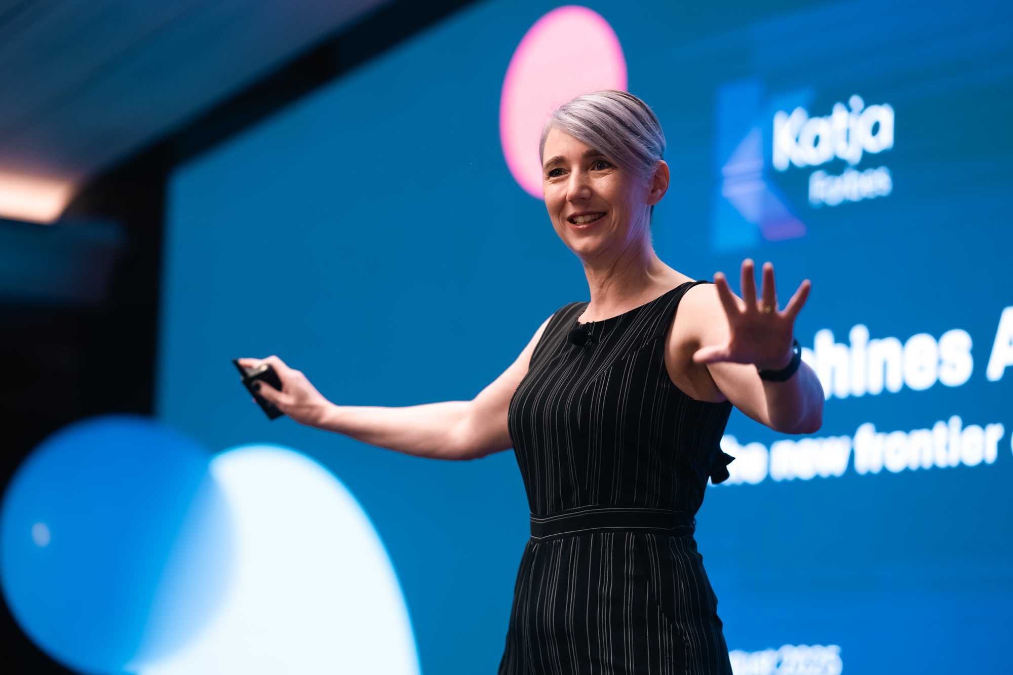 Katja Forbes delivering a keynote on a dark stage lit in blue and pink