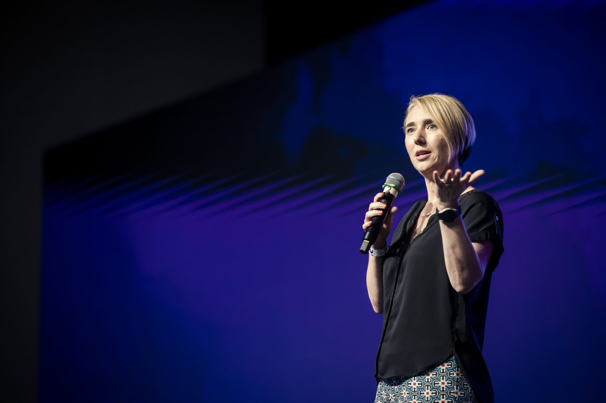 Katja Forbes speaking with a microphone against a deep blue and purple stage wash